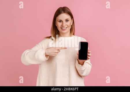 Porträt einer zufriedenen Frau, die das Smartphone in den Händen hält, auf ein leeres mobiles Display mit Platz für Werbung zeigt und einen weißen Pullover trägt. Innenaufnahme des Studios isoliert auf rosa Hintergrund. Stockfoto