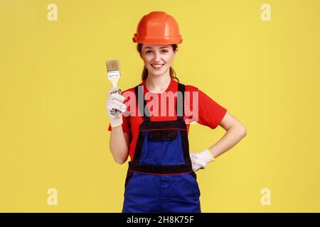 Positive junge Erwachsene Baumeisterin Frau, die mit Pinsel in den Händen steht, Arbeitsuniform und Schutzhelm trägt, hält die Hand auf der Taille. Innenaufnahme des Studios isoliert auf gelbem Hintergrund. Stockfoto