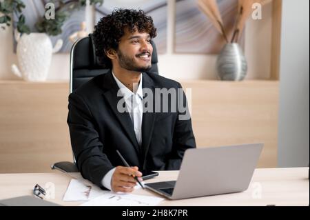 Indischer positiver Geschäftsmann, in formeller Kleidung, sitzt am Tisch im Büro, während er in einem Laptop arbeitet, positiv beiseite schaut, über ein Startup nachdenkt oder von einem zukünftigen Urlaub träumt Stockfoto