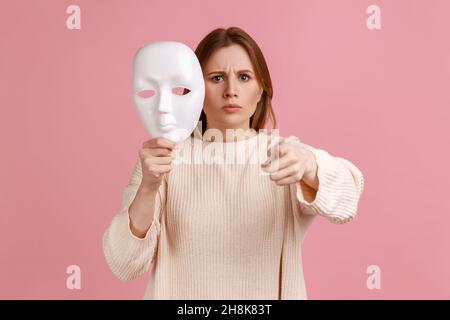 Portrait einer streng herrisch blonden Frau, die mit dem Finger auf die Kamera zeigt und weiße Maske in der Hand hält, will die Persönlichkeit verändern und trägt einen weißen Pullover. Innenaufnahme des Studios isoliert auf rosa Hintergrund. Stockfoto