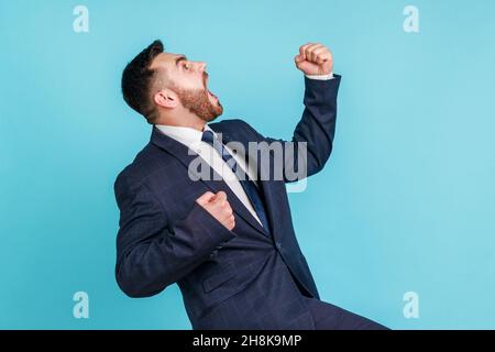 Seitenansicht eines überglücklich aussehenden Mannes, der einen Anzug im offiziellen Stil trägt, der mit erhobenen Fäusten und Schreien eine siegreiche Geste zum Ausdruck bringt und den Sieg feiert. Innenaufnahme des Studios isoliert auf blauem Hintergrund. Stockfoto