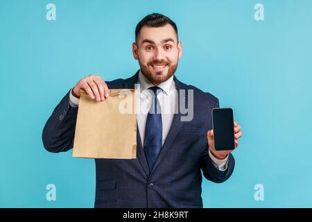 Glücklicher Mann, der einen offiziellen Anzug trägt, der ein leeres Display mit Handy und einer Öko-Papiertasche hält, Online-Shopping, schnelle Lieferung, Marktplatz. Innenaufnahme des Studios isoliert auf blauem Hintergrund. Stockfoto