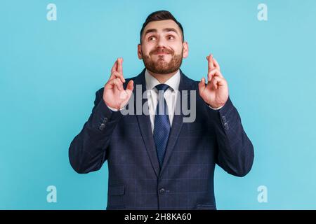 Porträt eines hoffnungsvollen Mannes mit Bart, der einen Anzug im offiziellen Stil trägt, der die Finger kreuzt, um Glück zu haben, einen Wunsch zu machen, vom Innersten zu träumen, ein Ritual. Innenaufnahme des Studios isoliert auf blauem Hintergrund. Stockfoto