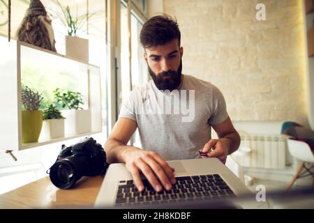 Junger bärtiger Mann, der eine SD-Karte einsteckt, um Fotos mit seiner Digitalkamera und seinem Laptop zu überprüfen. Stockfoto