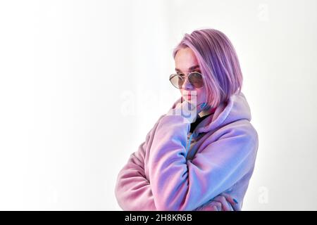 Serious junge Frau in Hoodie und stilvolle Sonnenbrille berühren Kinn und denken über die Entscheidung auf weißem Hintergrund Stockfoto