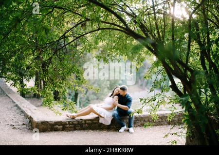 Der Mann umarmt die Frau, während er auf einer Steingrenze im grünen Park sitzt Stockfoto