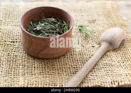 Rosmarin in einer Holzschüssel mit einem Löffel auf dem Tisch trocknen. Rustikaler Stil. Das trockene Gewürz wird zu Fleisch, Salaten, Suppen oder Getränken hinzugefügt. Zutat für den bou Stockfoto