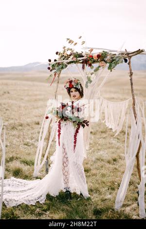 Neben dem Hochzeitbogen steht die Braut in einem Korbkleid und einem Kranz mit einem Blumenstrauß Stockfoto