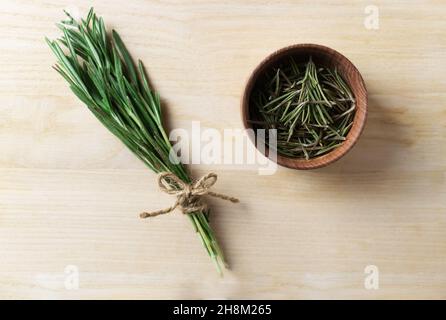 Ein Bund frischer Rosmarin, gebunden mit einer Jute, trocknet in einer Holzpiae auf einem Holztisch. Rustikaler Stil. Das Gewürz wird zu Fleisch, Salaten, Suppen oder Getränken hinzugefügt. Stockfoto