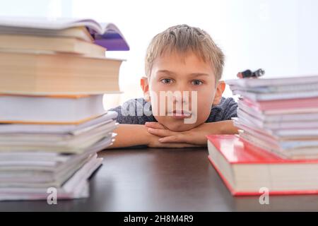 Teenager, der zu Hause Hausaufgaben macht. Das Kind sitzt auf seinem Schreibtisch zwischen einem Haufen von Büchern, Lehrbüchern und Schulübungsbüchern Stockfoto