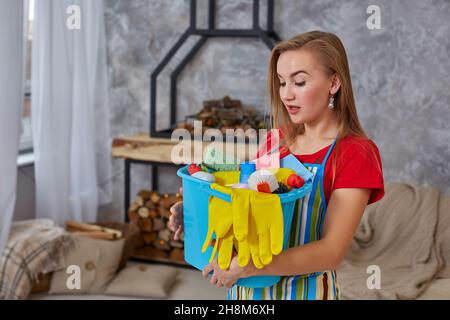 Eine Reinigungsfrau steht im Wohnzimmer und hält einen blauen Eimer mit Chemikalien und Einrichtungen zum Aufräumen in der Hand Stockfoto