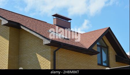 Eine Nahaufnahme eines Backsteinhauses mit einem braunen Asphaltschindeldach, Soffit und Faszien, Dachfenster, Kamin, Dachfenster und Dachrinnen-System mit Stockfoto