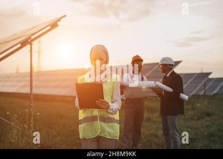 Die muslimische Frau hijab schrieb auf die Zwischenablage, während ihre männlichen Kollegen hinter Helmen standen und Blaupausen untersuchten. Multirassische Menschen, die die Produktion von Sonnenenergie entwickeln. Stockfoto