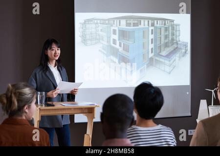 Asiatische junge Frau, die Bericht vorstellt, während sie in der Nähe des großen Monitors mit Bauplan und Menschen steht, die ihr zuhören Stockfoto