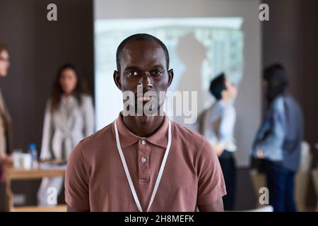 Porträt eines jungen afrikanischen Geschäftsmannes, der die Kamera anschaut, während er im Sitzungssaal mit anderen Menschen im Hintergrund steht Stockfoto