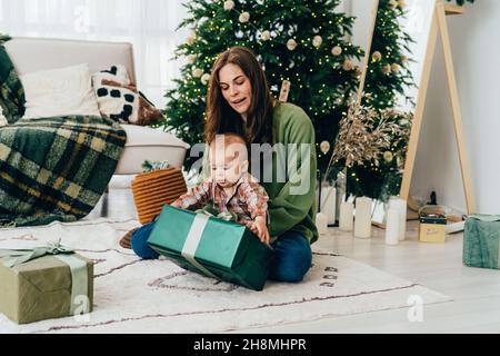 Junge kaukasische verschiedene Rothaarige Mumie sitzt auf dem Boden mit Kleinkind auspackert Weihnachtsgeschenk. Neujahrskonzept. Familientraditionen des Schenkens. Stockfoto