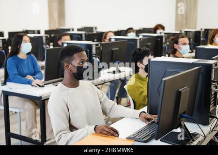 Multirassische junge Studenten, die während des Coronavirus-Ausbruchs im Klassenraum Computer mit Sicherheitsmasken benutzen - Focus african Boy Face Stockfoto