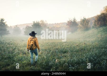 Wandern in herbstlicher Natur. Frau mit Rucksack und Hut, die am nebligen Morgen im Gras herumläuft. Touristen genießen Herbstsaison Stockfoto