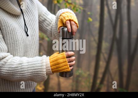 Wanderer öffnen Thermoskannen mit heißem Getränk im Wald. Frau mit Strickpullover und fingerlosen Wollhandschuhen. Erfrischung während der Wanderung im Freien Stockfoto