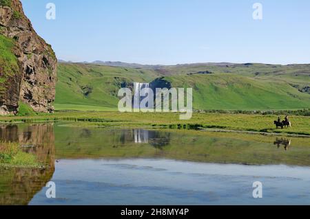 Wassermassen stürzen senkrecht in die Tiefe, Spiegelung im Wasser, grüne Landschaft, zwei Reiter auf isländischen Pferden, Skogafoss, Island Stockfoto