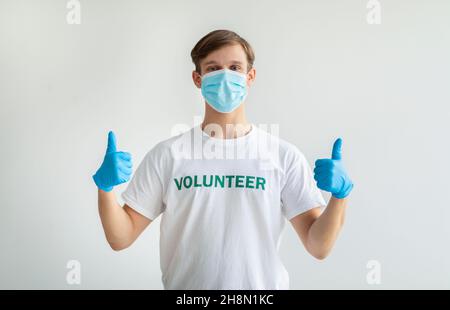 Wohltätigkeitskonzept. Millennial-Mann in weißem Freiwilligen-T-Shirt zeigt Daumen nach oben, trägt medizinische Maske und Handschuhe Stockfoto
