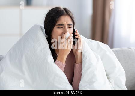 Verzweifelte, in eine Decke gewickelte junge indische Frau bläst Nase in Serviette, niesen, telefoniert auf der Couch Stockfoto