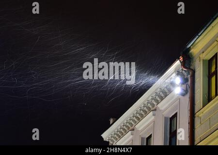 Ein Lichtstrahl aus einer unter dem Dach montierten Lampe beleuchtet die wirbelnden Schneeflocken an einem Winterabend. Schnee. Stockfoto