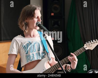 Junge leidenschaftliche Musikerin, die im Studio Gitarre spielt und während der Probe singt. Rockmusikerin, die neben einem Mikrofon steht Stockfoto