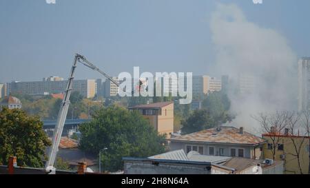 Feuerwehrleute auf dem Plattformwagen, die auf das Dach des Gebäudes fahren, um den Brand in der Stadt zu löschen. Feuerwehrleute im Haus versuchen, Flammen mit Wasser aus der Abteilung zu löschen, helfen mit Smog und Schutz Stockfoto