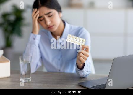 Verzweifelte, traurige junge indische Dame, die am Arbeitsplatz an Schmerzen, Migräne und Krankheiten leidet, hält Pillen in der Tablette Stockfoto