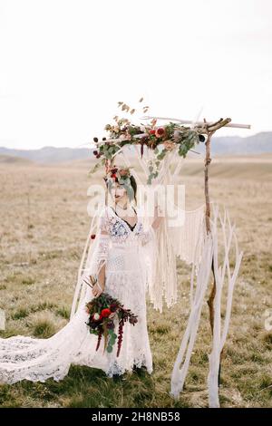 Braut in einem weißen Korbkleid und einem Kranz steht auf einem Feld neben einem Hochzeitbogen Stockfoto