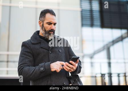 Porträt eines Geschäftsmannes in schwarzer Kleidung, der vor dem Firmengebäude steht und auf das Smartphone schaut. Schwarzer bärtiger Mann verwendet Smartphone. Stockfoto