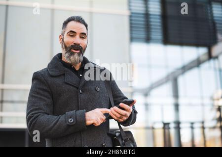 Porträt eines glücklichen Geschäftsmannes in schwarzer Kleidung, der vor dem Firmengebäude steht. Schwarzer bärtiger Mann genießt es, Smartphone zu benutzen. Stockfoto