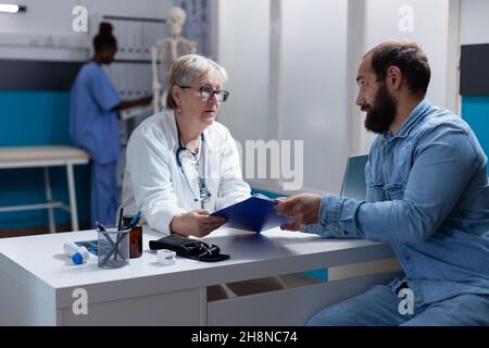 Ärztin gibt dem Patienten die Zwischenablage, um die Einwilligung zur Behandlung zu unterzeichnen. Arzt bittet den Mann um Unterschrift auf den Kontrollpapieren bei einem Arztbesuch, um die Krankheit zu heilen. Person, die Untersuchungsdateien signiert. Stockfoto