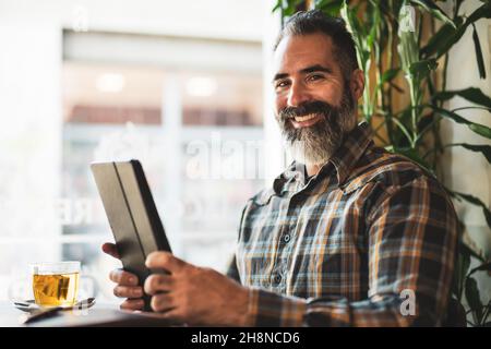 Der Erwachsene Mann sitzt im Café und benutzt ein digitales Tablet. Er ist entspannend. Stockfoto