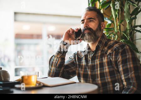 Der Erwachsene Mann sitzt im Café und macht seinen täglichen Zeitplan. Er telefoniert und benutzt Notizblock. Stockfoto