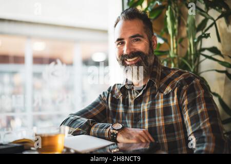 Der Erwachsene Mann sitzt im Café und macht seinen täglichen Zeitplan. Er blickt auf die Kamera und lächelt. Stockfoto