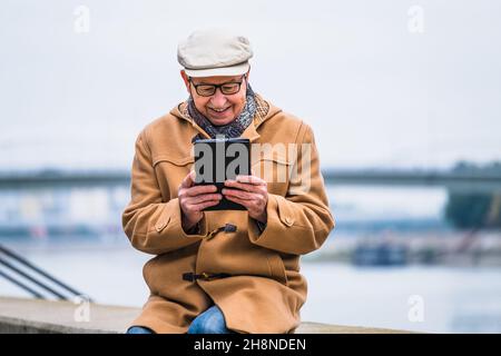 Porträt eines fröhlichen älteren Mannes im Wintermantel, der ein digitales Tablet verwendet. Stockfoto