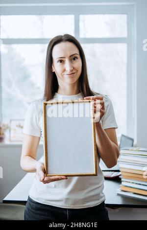 Rahmenbild, Plakat, Diplom, Urkunde in weiblichen Händen. Junge Brünette Frau hält leeren leeren hölzernen Fotorahmen auf Tisch mit Laptop Stockfoto