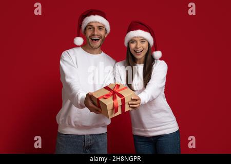 Frohe Feiertage. Fröhliche Junge Ehegatten Geben Weihnachtsgeschenk-Box An Der Kamera Stockfoto