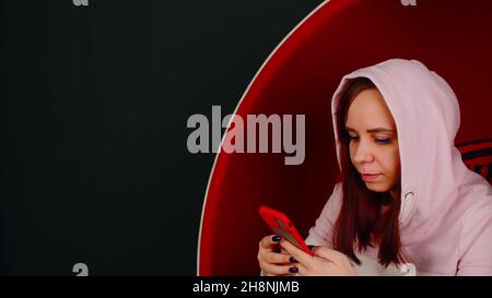 Junge Frau mit Mobiltelefon sitzt in einem Kugelstuhl auf schwarzem Hintergrund. Weibliche Browsing Smartphone und Ruhe in modernen Sessel Stockfoto