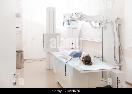 Arzt, der Röntgenaufnahme eines Patienten, der auf einer Liege lag. Radiologiezimmer des Krankenhauses. Techniker, der ein Röntgengerät anpasst. Weibchen, die auf einem Bett liegt, scannt Stockfoto