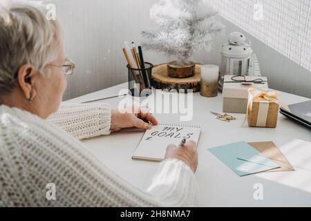 Gesunde Neujahrvorsätze für ältere Erwachsene. Ältere ältere alte Frau in weißem Pullover schreiben handschriftlichen Text My Goals in offenen Notizblock auf dem Stockfoto