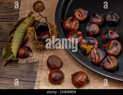 Geröstete Kastanien und Kastanien, eine davon mit Igel, auf einem Backblech und auf einem Holztisch Stockfoto
