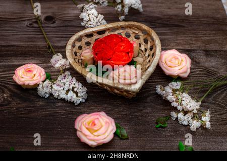 Schokolade blüht in einem Korb in Form eines Herzens. Rosen, Pfingstrosen auf braunem Holzhintergrund. Stockfoto