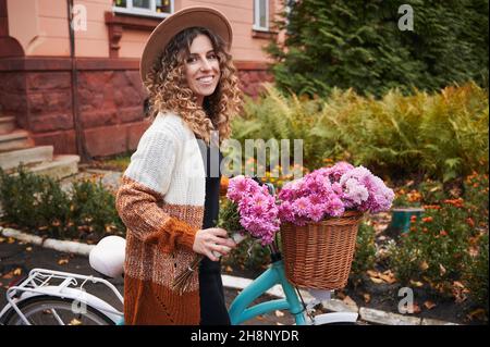 Hübsche Frau mit lockigen Haaren, die auf die Kamera schaut und lächelt, während sie das Fahrrad mit Blumen im Korb hält. Fröhliche junge Frau mit elegantem Hut und Strickjacke, die Zeit im Freien verbringt. Stockfoto