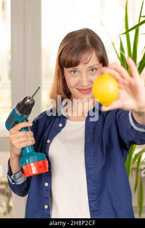 Eine junge hübsche Frau in einem blauen Hemd hält einen Schraubendreher mit einem Bohrer und einer hellen Zitrone vor dem Hintergrund eines hellen Fensters und grünen Blättern Stockfoto