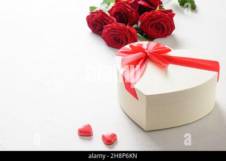 Valentinstag-Geschenkbox als Herz, rote Schokoladenbonbons und Rosenstrauß auf weißem Nacktboden mit Kopierfläche. Nahaufnahme. Stockfoto