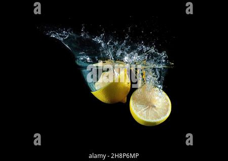 Zwei Hälften frischer Zitronen sinken tief in Wasser mit Spritzern vor schwarzem Hintergrund Stockfoto