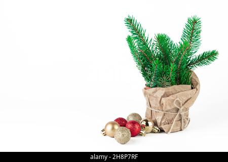 Kleiner, undekorierter Weihnachtsbaum in einem Topf, eingewickelt in Bastelpapier und Weihnachtsbaumkugeln auf weißem Hintergrund. Stockfoto